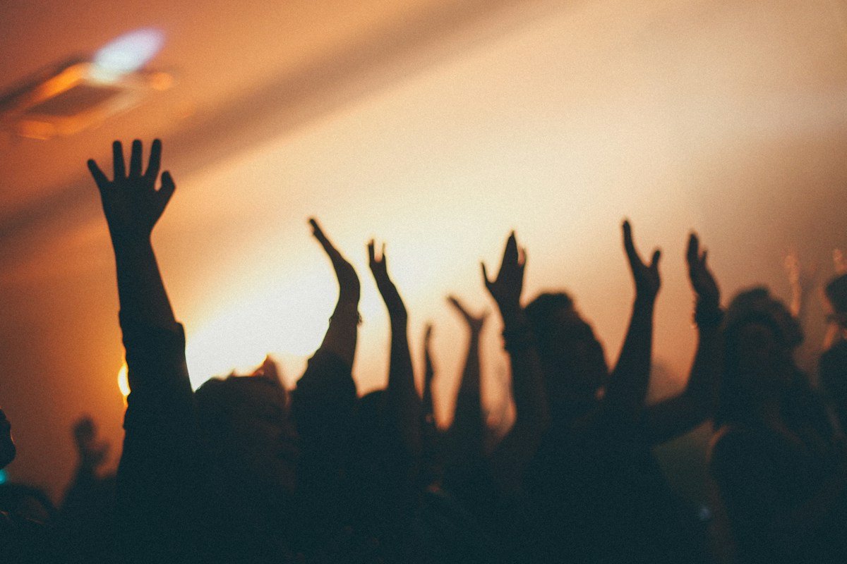 Silhouettes de bras levés dans la foule d’un concert, devant un faisceau de lumière orangé